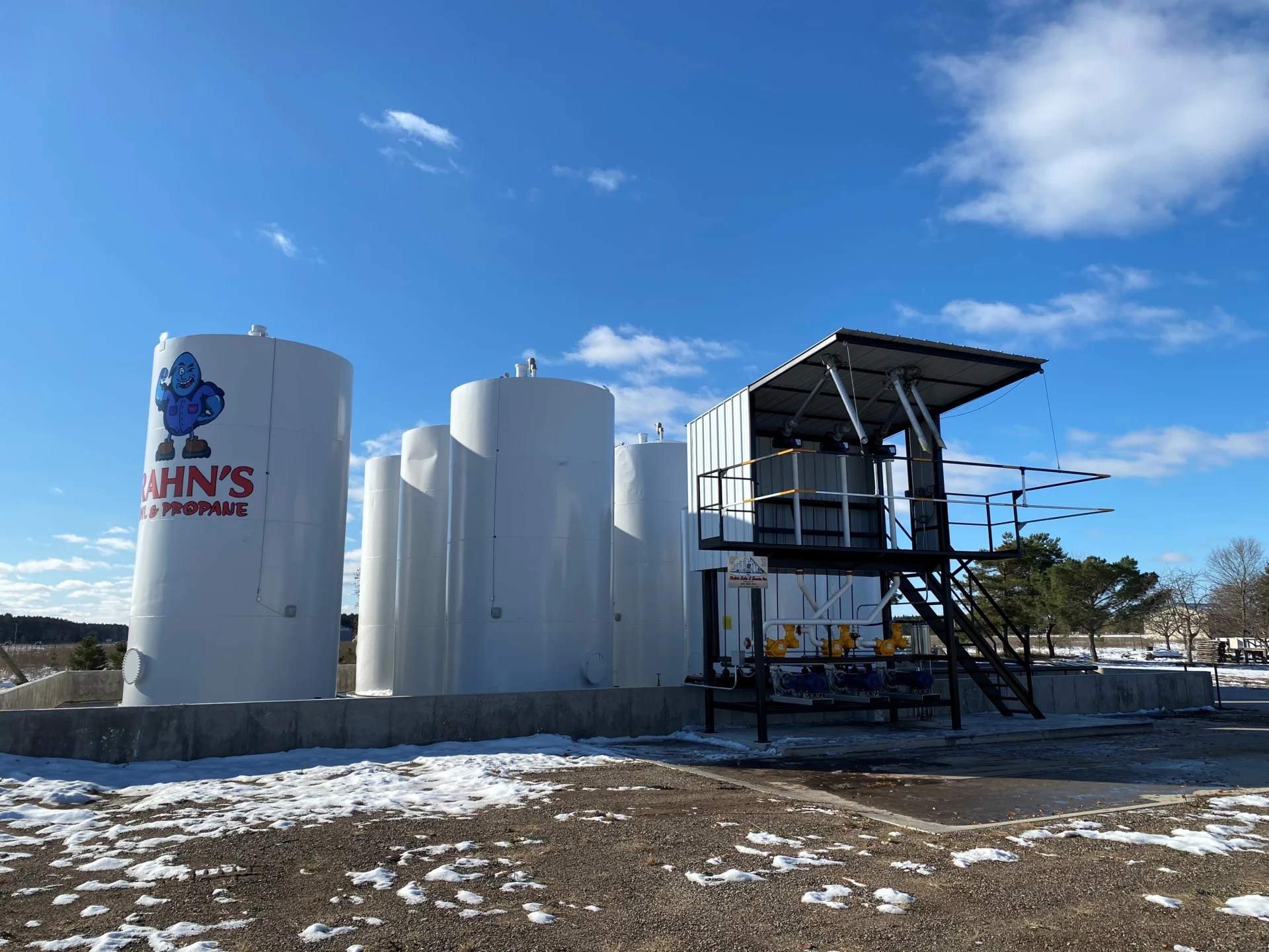 Four large white propane storage tanks and a small metal control building stand on a snowy lot under a blue sky. One tank displays "Hahn's Propane" and a cartoon mascot, highlighting expert Fuel System Installation & Repair in Minnesota. Trees and clouds in back.