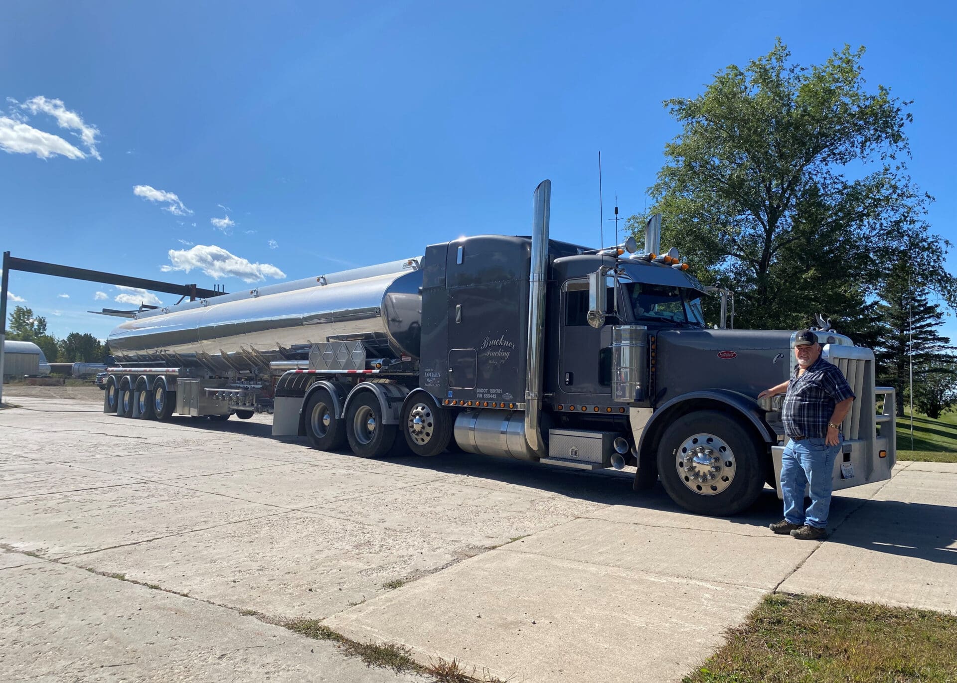 A man in a plaid shirt and cap stands beside a large, shiny black bulk fuel semi-truck with a long tanker trailer, parked on a concrete surface under a clear blue sky with some trees in the background.