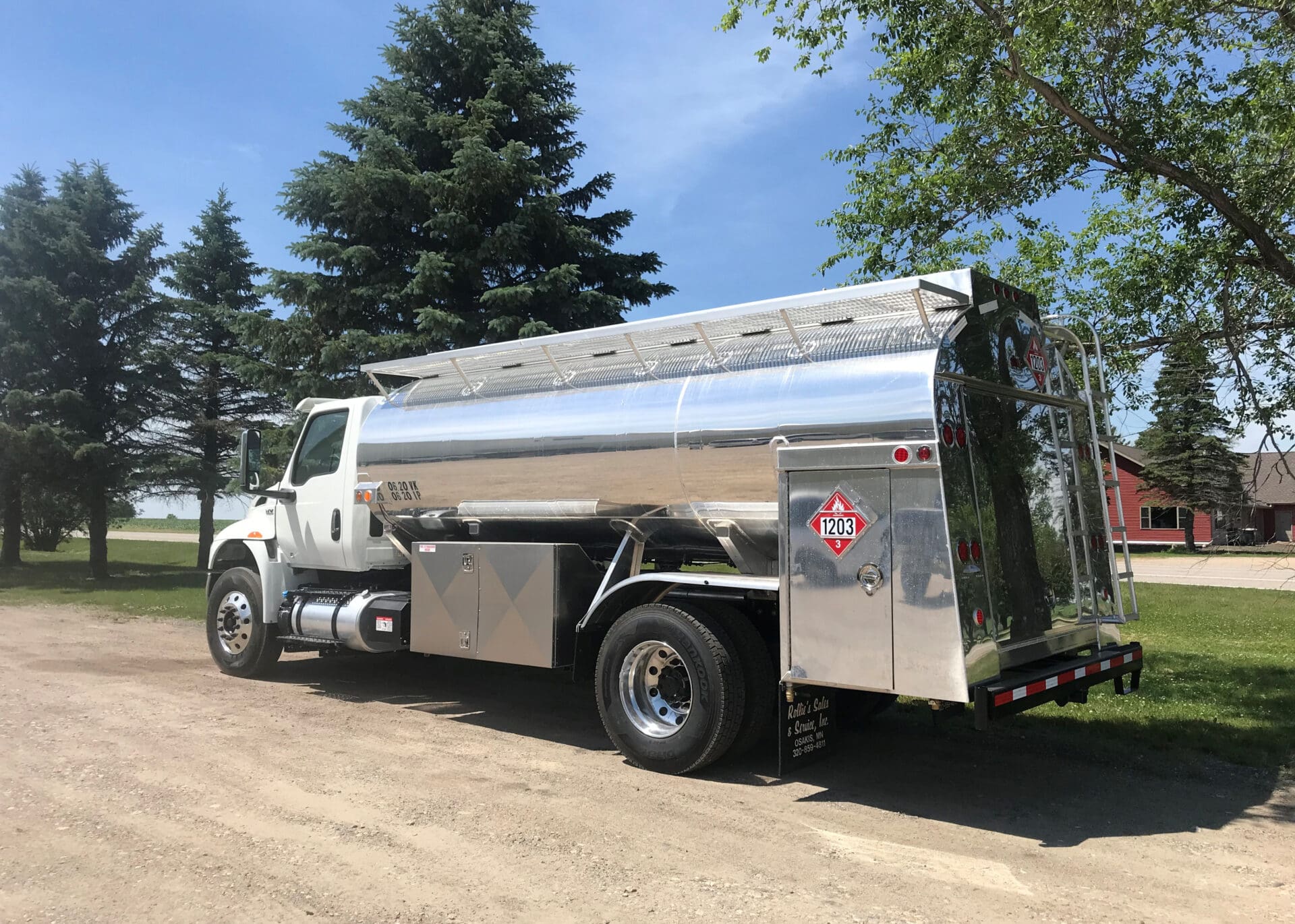 A shiny silver fuel delivery truck is parked on a dirt road beside green grass and trees under a clear blue sky, with a building visible in the background.