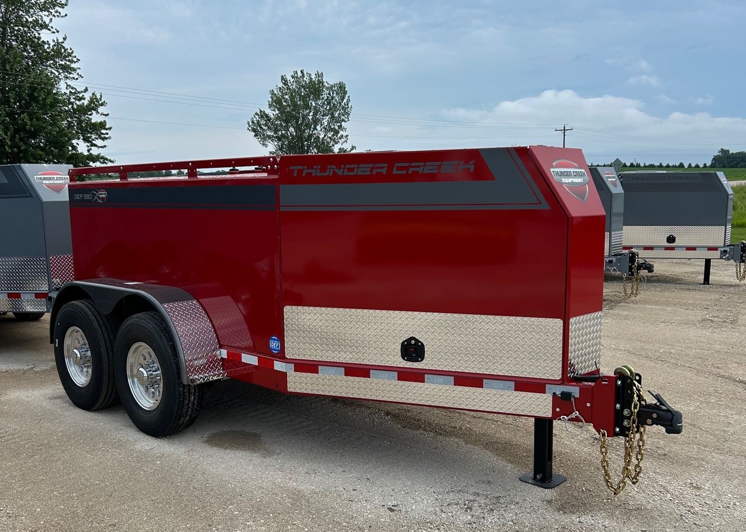 A red Thunder Creek utility trailer with two axles and chrome accents is parked on a gravel lot, showcasing the quality of Thunder Creek fuel trailers in Minnesota, with similar models visible in the background.